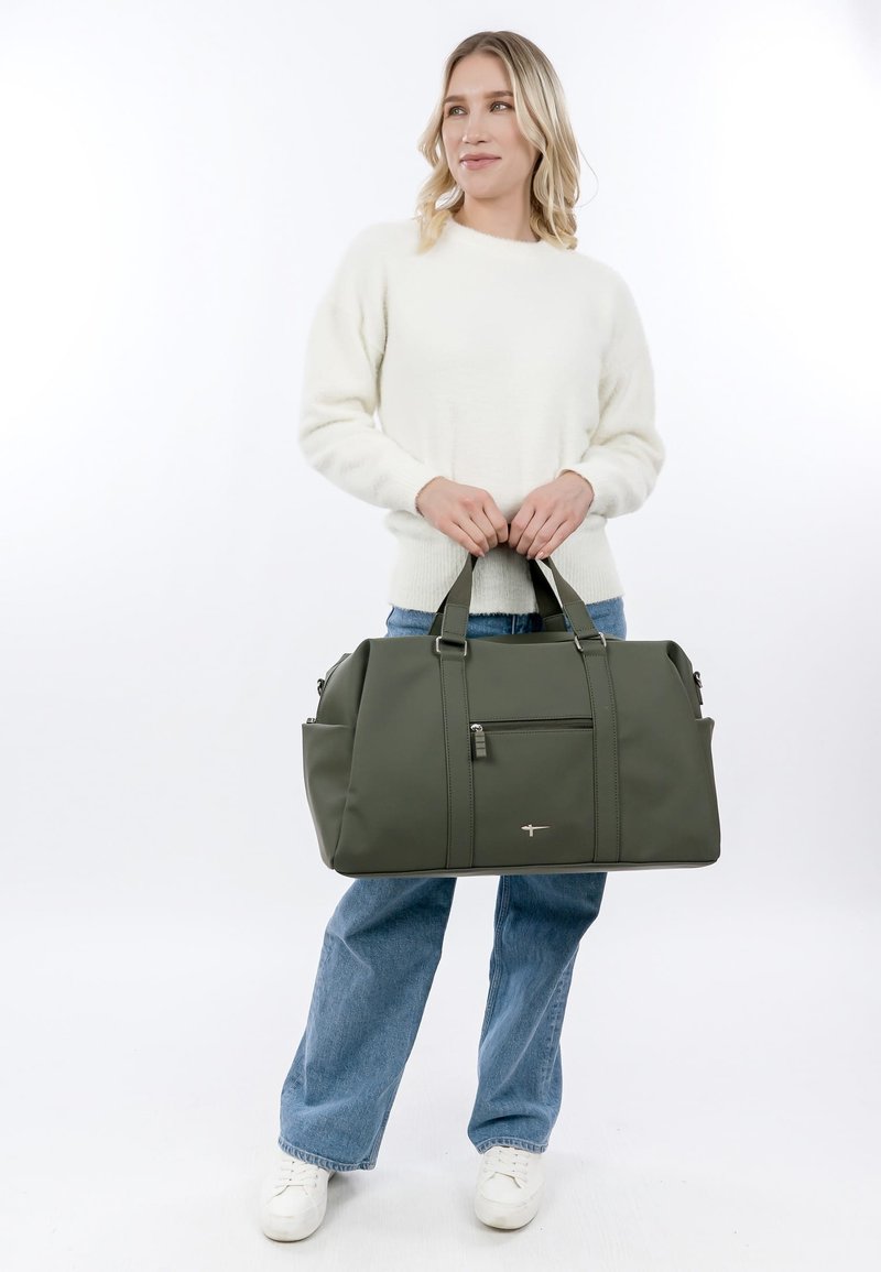 Vrouw in witte trui en spijkerbroek met grote olijfgroene reistas met voorvak met ritssluiting.