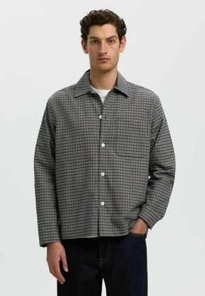 Homme aux cheveux courts et bouclés portant une chemise grise à carreaux boutonnée sur un haut blanc et un jean foncé, debout avec une main dans la poche.