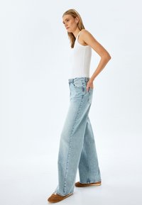 Jeans de mezclilla de pierna ancha en azul claro con cintura alta, que cuentan con cinco bolsillos y un ajuste relajado. Combinados con una camiseta sin mangas blanca ajustada y zapatos marrones.