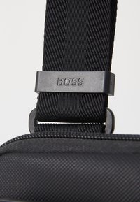 Black fabric bag strap with textured finish, featuring a matte black hardware adjustment piece engraved with "BOSS." Smooth zipper detail visible.