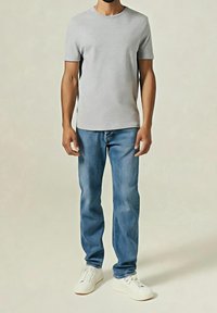 Homme portant un t-shirt gris clair à manches courtes, un jean bleu et des baskets blanches, debout devant un fond beige uni.
