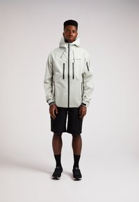 Giacca impermeabile grigia chiara con capuccio, tasche con zip nere e dettagli del logo. Indossata sopra pantaloni cargo neri e calze nere.