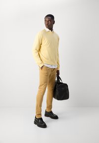 Helle gelbe Pullover über einem weißen Hemd mit Kragen, beige Slim-Fit-Hose und schwarze Schuhe. Er hält eine schwarze Tasche mit abgerundeter Form und kurzen Griffen.