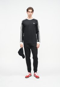 Melna krekla ar baltu apkakli un trīs baltām svītrām uz piedurknēm, kombinēts ar melnām biksēm un sarkanām sporta kurpēm. Melna cepure turēta rokā.