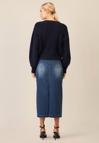 Marinefarbener gerippter Pullover mit weiten Ärmeln, kombiniert mit einem knielangen Jeansrock in verblasster Ausführung und mit Nietenverzierungen. Schwarze Riemchen-High-Heels.