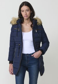 Giacca puffer blu navy con cappuccio in pelliccia sintetica, dotata di numerose tasche con cerniera, vita regolabile e dettagli in metallo dorato.