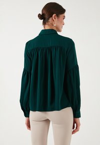 Green long-sleeve blouse in smooth fabric, featuring a pleated back design and buttoned cuffs. The collar is standard; styled for a relaxed fit.