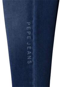 Pierna de jeans de mezclilla azul oscuro con un sutil logotipo vertical "PEPE JEANS" impreso en texto blanco desgastado a lo largo de la costura.