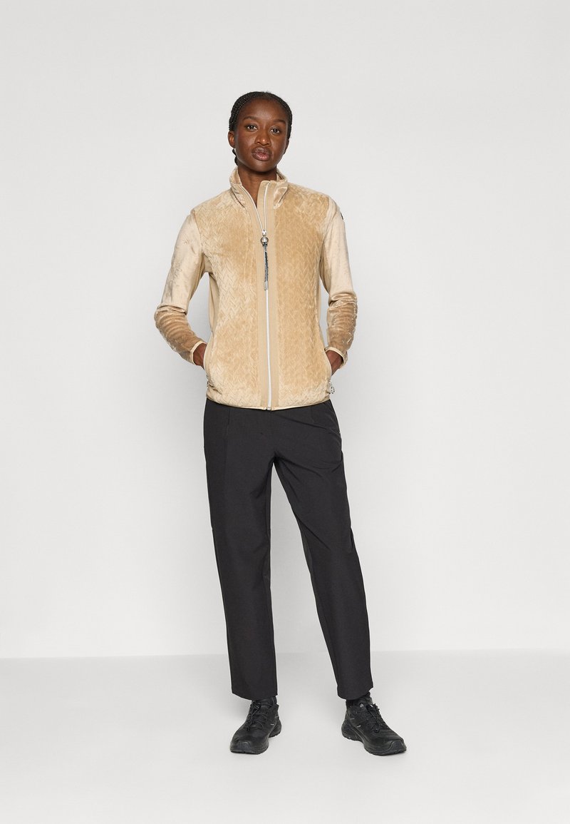 Beżowa kurtka zapinana na zamek z fakturowanym przodem, gładkimi rękawami i kołnierzem. Połączona z czarnymi spodniami i czarnymi butami, umiejscowiona na jednolitym tle.