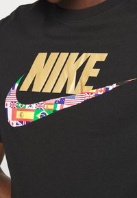 Svart bomull t-shirt med en stor gyllene "NIKE"-logotyp och en färgglad swoosh bestående av olika nationella flaggor.