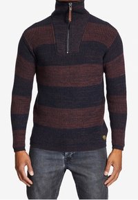 Uomo che indossa un maglione a coste con mezza zip, caratterizzato da ampie strisce orizzontali blu navy e marroni, abbinato a jeans grigi scuri.