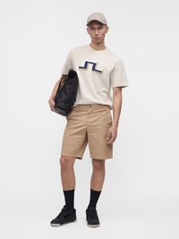 Jeune homme en T-shirt beige avec logo abstrait, short beige clair, baskets noires, chaussettes noires, casquette beige, tenant un grand sac noir, debout.
