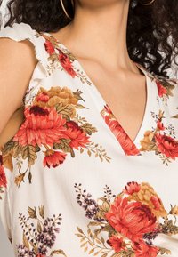 Blusa blanca con flores que presentan tonos rojos y dorados, escote en forma de V y detalles de volantes en los hombros. Tejido suave con un patrón delicado.