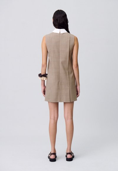 Modèle avec de longs cheveux, portant une robe sans manches en tartan marron avec un col blanc, vue de derrière, se tenant devant un fond uni.