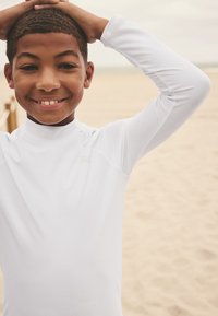 Vit långärmad rash guard med hög krage, bärs av ett leende barn. Slät textur med sydda sömmar. Sandig strand i bakgrunden.