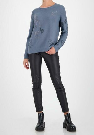 Mujer con un suéter azul claro con estampados, pantalones de cuero negros y botines negros, de pie con una mano en el bolsillo.
