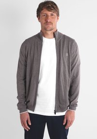Homme aux cheveux châtain clair portant un cardigan zippé gris sur un t-shirt blanc et un pantalon foncé, se tenant contre un fond uni clair.