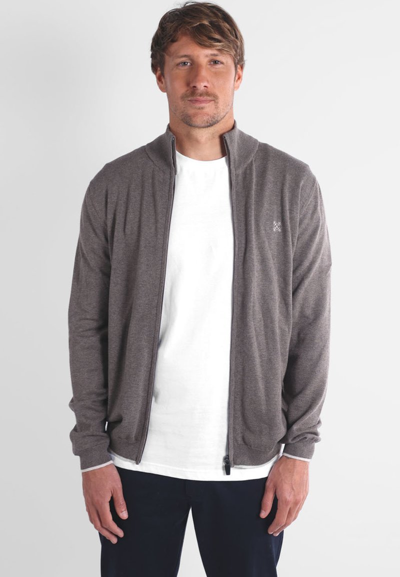 Homme aux cheveux châtain clair portant un cardigan zippé gris sur un t-shirt blanc et un pantalon foncé, se tenant contre un fond uni clair.