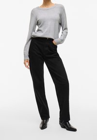 Personne portant un pull à manches longues gris clair, un pantalon noir taille haute et des bottines noires, debout avec une main dans la poche.