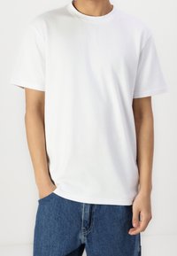 Personne portant un t-shirt blanc uni à manches courtes et un jean en denim bleu, avec une main dans la poche et l'autre détendue sur le côté, sur un fond uni.