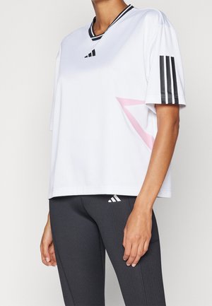 Personne portant un t-shirt de sport blanc à manches courtes avec des rayures noires sur les manches et un détail géométrique rose, associé à des leggings noirs côtelés.
