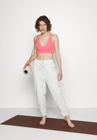 Top desportivo rosa, calças de jogging brancas largas com cordão, e um tapete de yoga enrolado segurado com uma mão, sobre um tapete de yoga castanho.