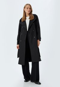 Czarny trench coat wykonany z gładkiego materiału, z szerokim kołnierzem, podwójnym zapięciem na guziki i dużymi guzikami. Nosi się go na białej bluzce i czarnych spodniach.