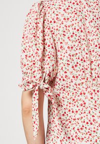 Ärmdetalj av vitt tyg med små röda blommönster, knutet i en rosett vid ärmlängd av puffärm.