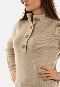 Maglione a coste beige con colletto alto, tre grandi bottoni dorati e maniche lunghe. Texture morbida, silhouette aderente e senza motivi.