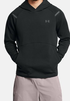 Osoba ubrana w czarną bluzę z kapturem Under Armour z przednimi kieszeniami oraz jasne spodnie, ręce wsunięte w kieszenie.