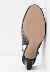 Chaussure à talon noire avec une tige texturée et embossée, et une semelle lisse de couleur claire. Présente un design ajouré et un petit talon carré.