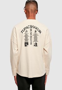 Cămașă crem cu mânecă lungă și croială lejeră. Are text și imagini negre pe spate, referindu-se la Tupac Shakur și versurile pieselor.