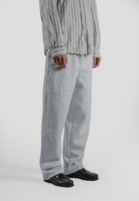 Osoba ubrana w luźne szare spodnie dresowe, czarne mokasyny oraz beżowo-białą pasiastą koszulę z długim rękawem na guziki na jednolitym tle.