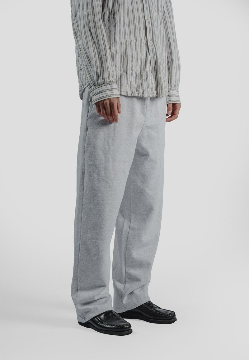 Osoba ubrana w luźne szare spodnie dresowe, czarne mokasyny oraz beżowo-białą pasiastą koszulę z długim rękawem na guziki na jednolitym tle.