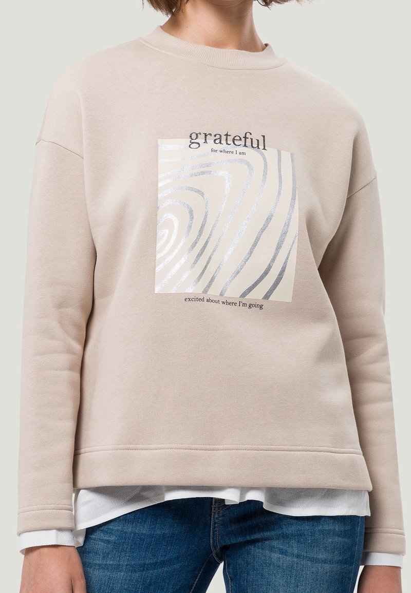 Beige sweatshirt met een ronde hals, uitgerust met een zilveromrand ontwerp en de tekst "dankbaar voor waar ik ben" en "enthousiast over waar ik naartoe ga."