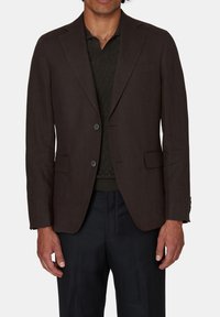 Blazer marrone su misura, monopetto con due bottoni, revers classici, due tasche frontali, indossato sopra una polo scura a trama e pantaloni scuri.