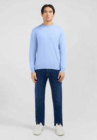 Pull en maille bleu avec des manches longues, un col rond, et un tissu texturé, assorti à un jean en denim foncé et des baskets blanches.
