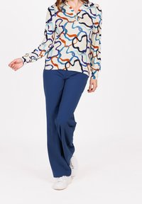 Valkoinen pitkähihaista paitaa, jossa on värikkäitä abstrakteja kuvioita sinisessä, oranssissa ja mustassa; yhdistetty tummansinisiin housuihin ja valkoisiin lenkkareihin.