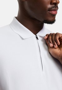 Weißes Poloshirt mit einem Viertel-Reißverschluss, das aus strukturiertem Stoff besteht und ein kontrastierendes schwarzes Reißverschlussdetail aufweist. Nahaufnahme des Kragens und einer Hand.