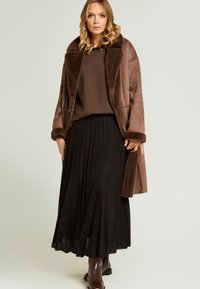 Abrigo marrón de piel sintética con un amplio cuello, combinado con una falda oscura plisada y botines marrones. Texturas suaves con colores contrastantes.