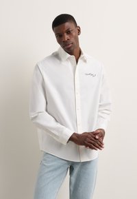 Camisa blanca de botones, hecha de una tela suave, con un cuello en punta y un detalle bordado negro en el pecho. Mangas de corte relajado.