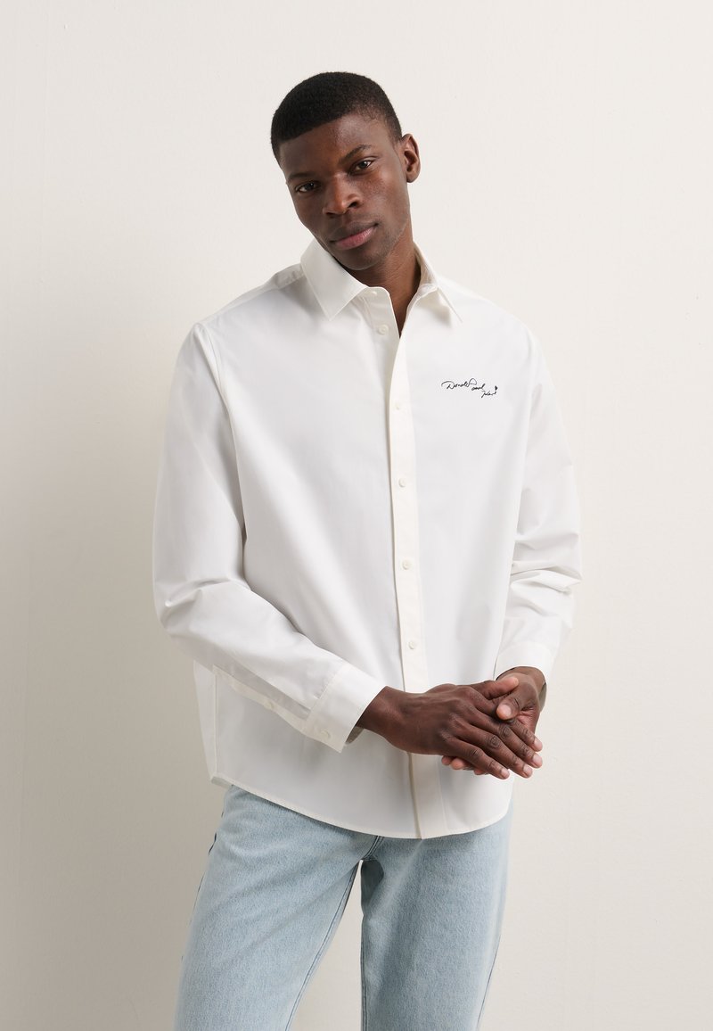 Camisa blanca de botones, hecha de una tela suave, con un cuello en punta y un detalle bordado negro en el pecho. Mangas de corte relajado.
