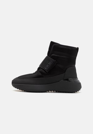 Botines negros con un upper suave, que presentan una correa de Velcro y acentos brillantes, suela de goma texturizada y diseño de punta redondeada.