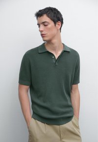 Polo a maniche corte di colore verde scuro in tessuto a maglia testurizzata, con colletto classico e placket a tre bottoni, abbinato a pantaloni beige.