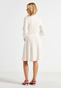 Lys cremefarvet sweaterkjole med lange ærmer, tætsiddende talje og flagrende nederdel. Blød tekstur med rynket kant. Sættes sammen med røde hæle sandaler.