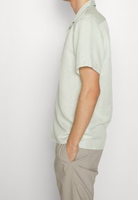 Camicia verde chiaro a maniche corte con un sottile motivo circolare, colletto abbottonato e vestibilità rilassata, abbinata a pantaloni beige.