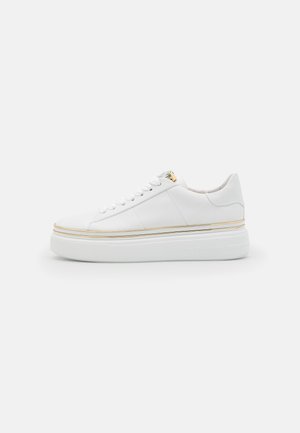Zapatilla blanca de caña baja con suela gruesa, detalle de ribete dorado alrededor de la suela, cordones blancos y acentos mínimos en las costuras sobre la parte superior de cuero liso.