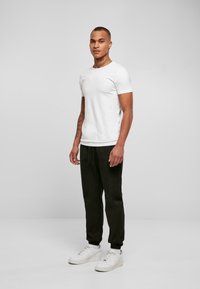 Vit bomull t-shirt, svarta svettbyxor och vita sneakers. Modellen står mot en plain grå bakgrund, med armarna avslappnat vid sidorna.