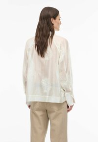 Blouse légère et semi-transparente de couleur blanc cassé avec broderie florale dans le dos. Caractérisée par une coupe ample et des manches longues, associée à un pantalon beige.