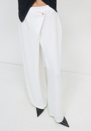 White wide-leg trousers featuring a wrap design with a front button closure. Fabric has a smooth texture. Paired with black pointed heels.